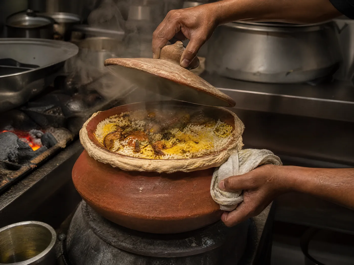A cook lifts the lid from a dough-sealed clay pot of biryani as steam escapes.